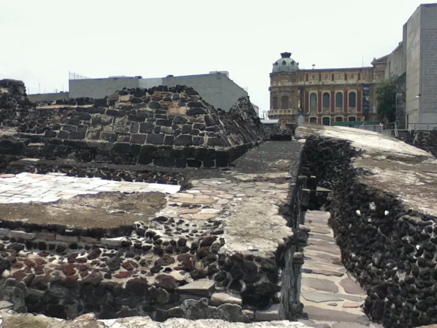 Ruínas do Templo Mayor asteca expostas no centro histórico de Cidade do México, com paredes de tezontle vermelho-escuro e edifícios coloniais ao fundo