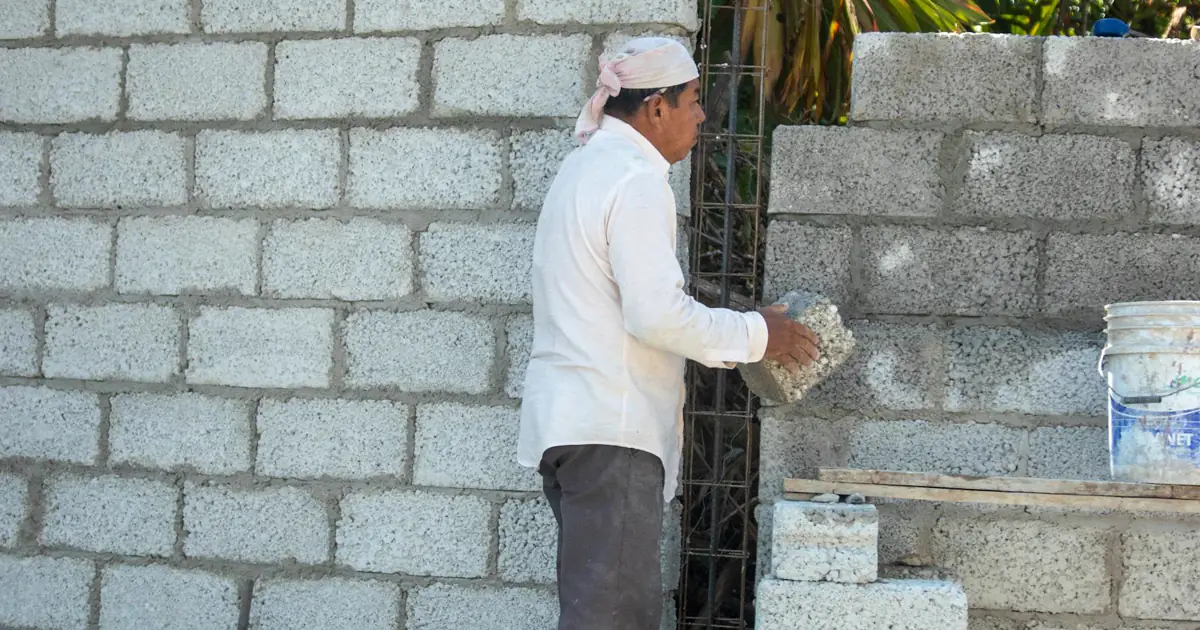 Pedreiro assentando blocos de concreto vazado com ferragem vertical em obra de alvenaria — escolha do elemento define resistência, peso e custo