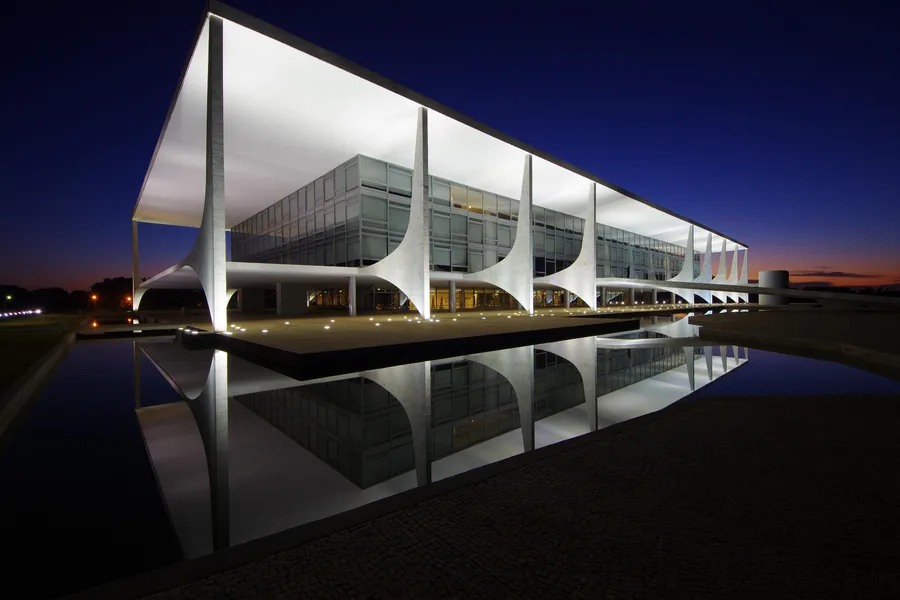 Fachada do Palácio do Planalto ao entardecer: pilares-asa de concreto branco iluminados refletidos no espelho d'água