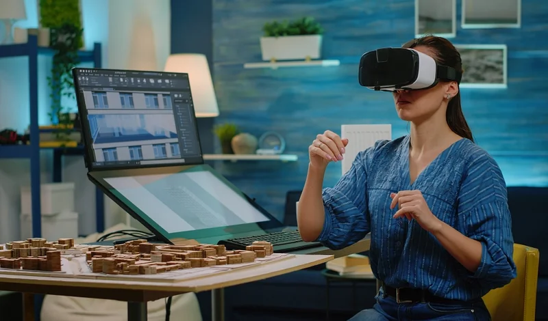 Arquiteta com headset de realidade virtual revisando modelo de edifício ao lado de maquete física e monitor com CAD
